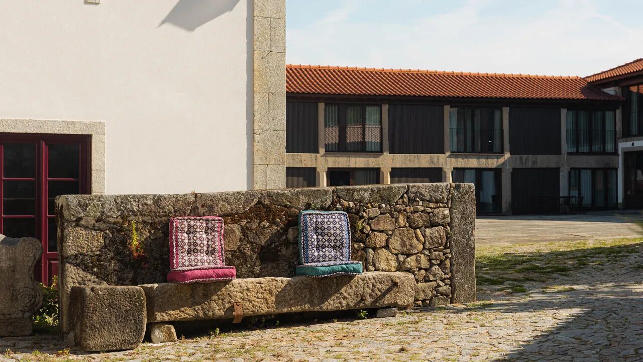 A stone bench with blankets in front of a building with a red-tiled roof and a stone wall.