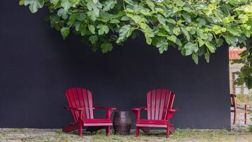 Two red Adirondack chairs and a barrel against a dark wall with green foliage overhead.