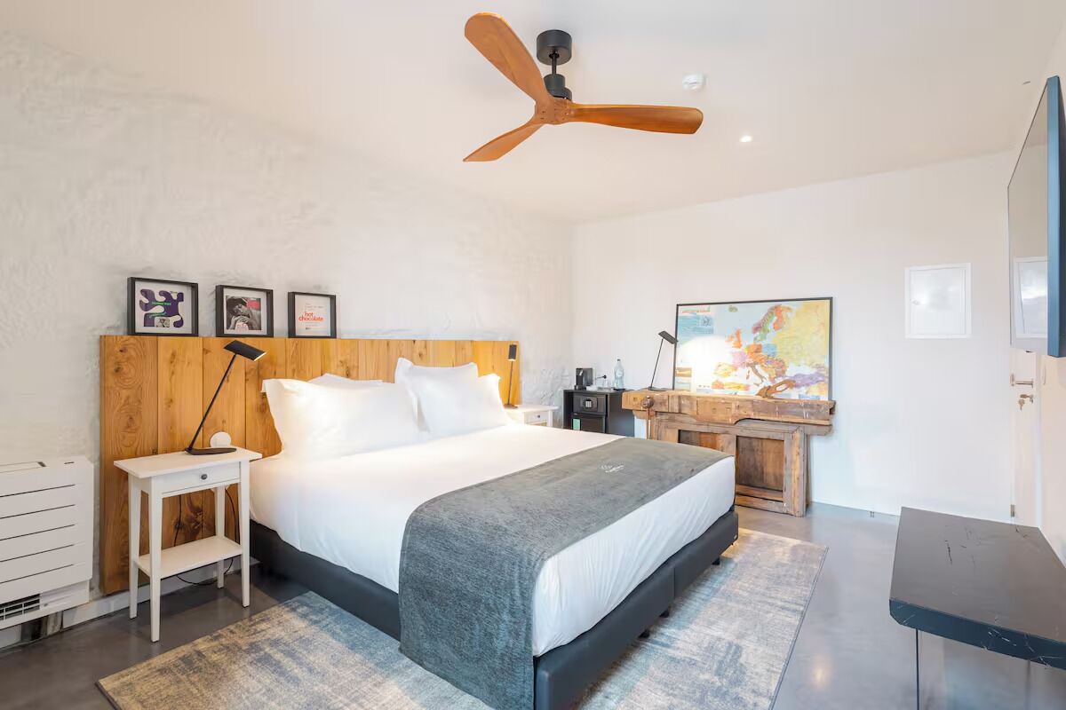 Un dormitorio moderno con cabecera de madera, cama con ropa blanca, escritorio con silla y ventilador de techo.