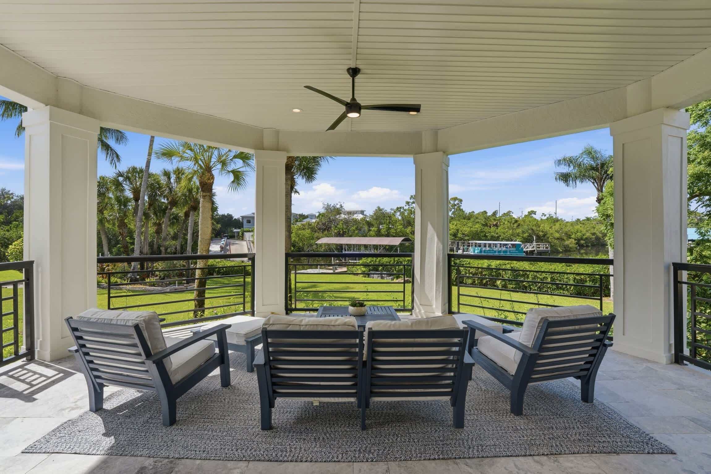 A covered outdoor seating area with a ceiling fan, a table with chairs, and a view of a well-maintained lawn and distant buildings.