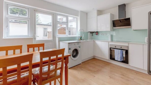 A modern kitchen with a washing machine, oven, and dining area with chairs. - Large Modern 4 BDR Home Near ExCel London (Greater London)