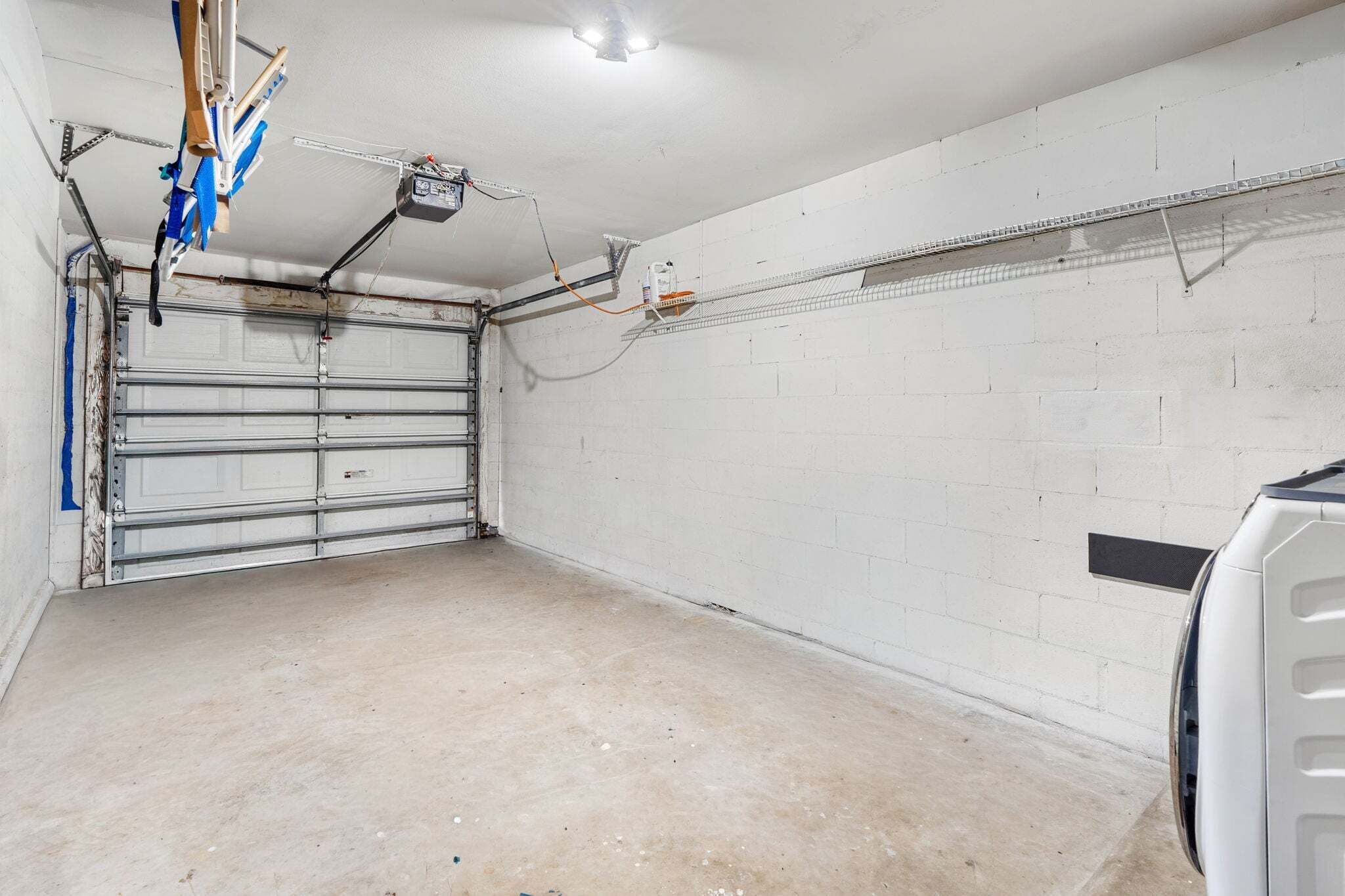 Un garaje con puerta cerrada, piso de concreto y pared blanca con estante metálico.