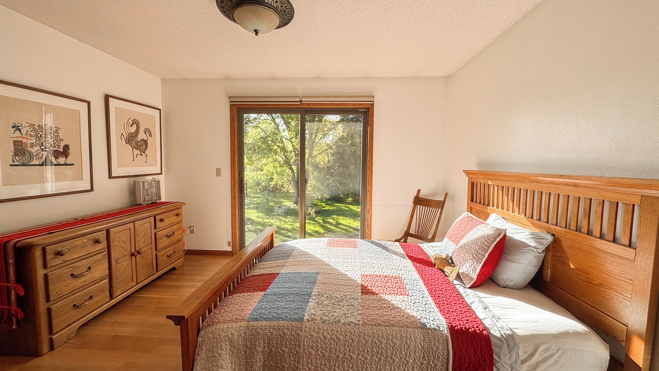 A bedroom with a wooden bed, a wooden bench, a dresser, and a large window with a view of greenery.