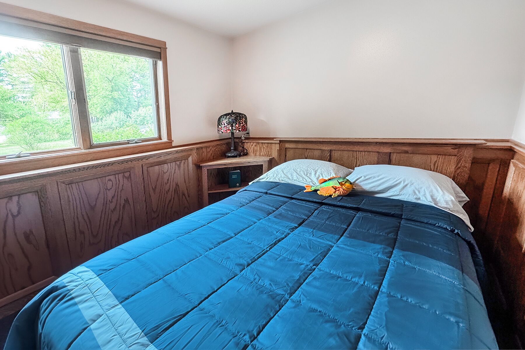 Une chambre avec une grande fenêtre, un lit recouvert d’une couette bleue et une table de chevet avec une lampe.