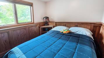 Une chambre avec une grande fenêtre, un lit recouvert d’une couette bleue et une table de chevet avec une lampe.