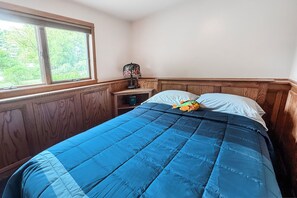A bedroom with a large window, a bed with a blue quilt, and a nightstand with a lamp.