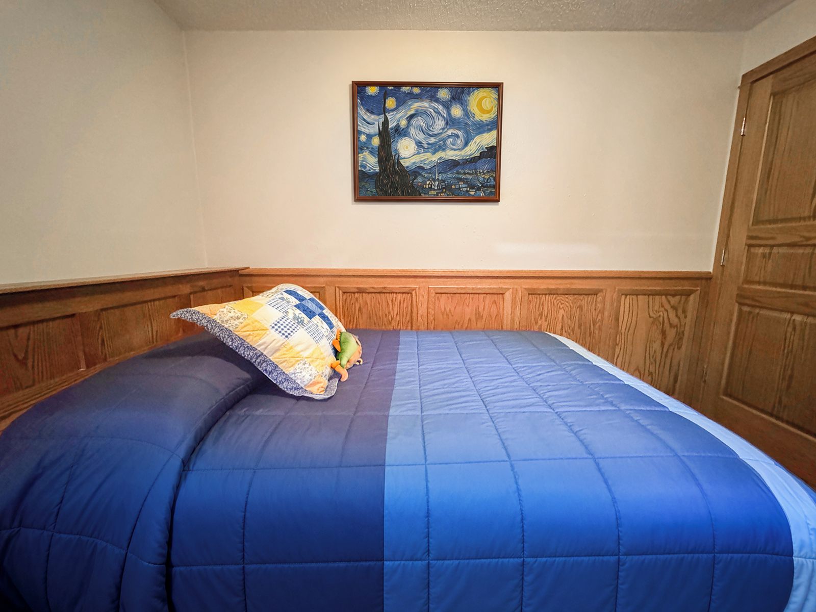 A bedroom with a blue bedspread, a quilted pillow, a painting of a night sky on the wall, and wooden paneling on the walls.