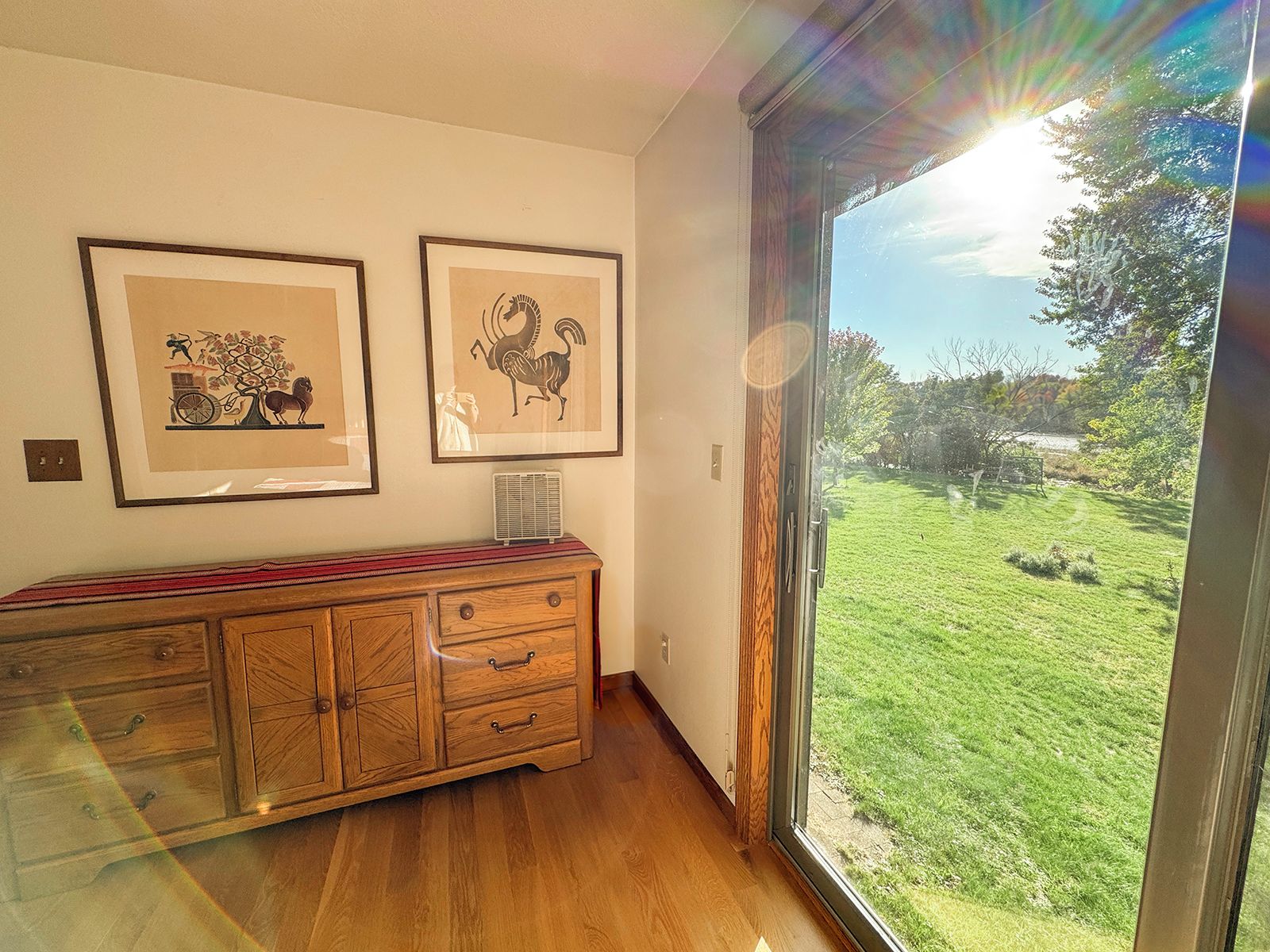 A room with a wooden cabinet, two framed artworks, and a large window with a view of a grassy area.