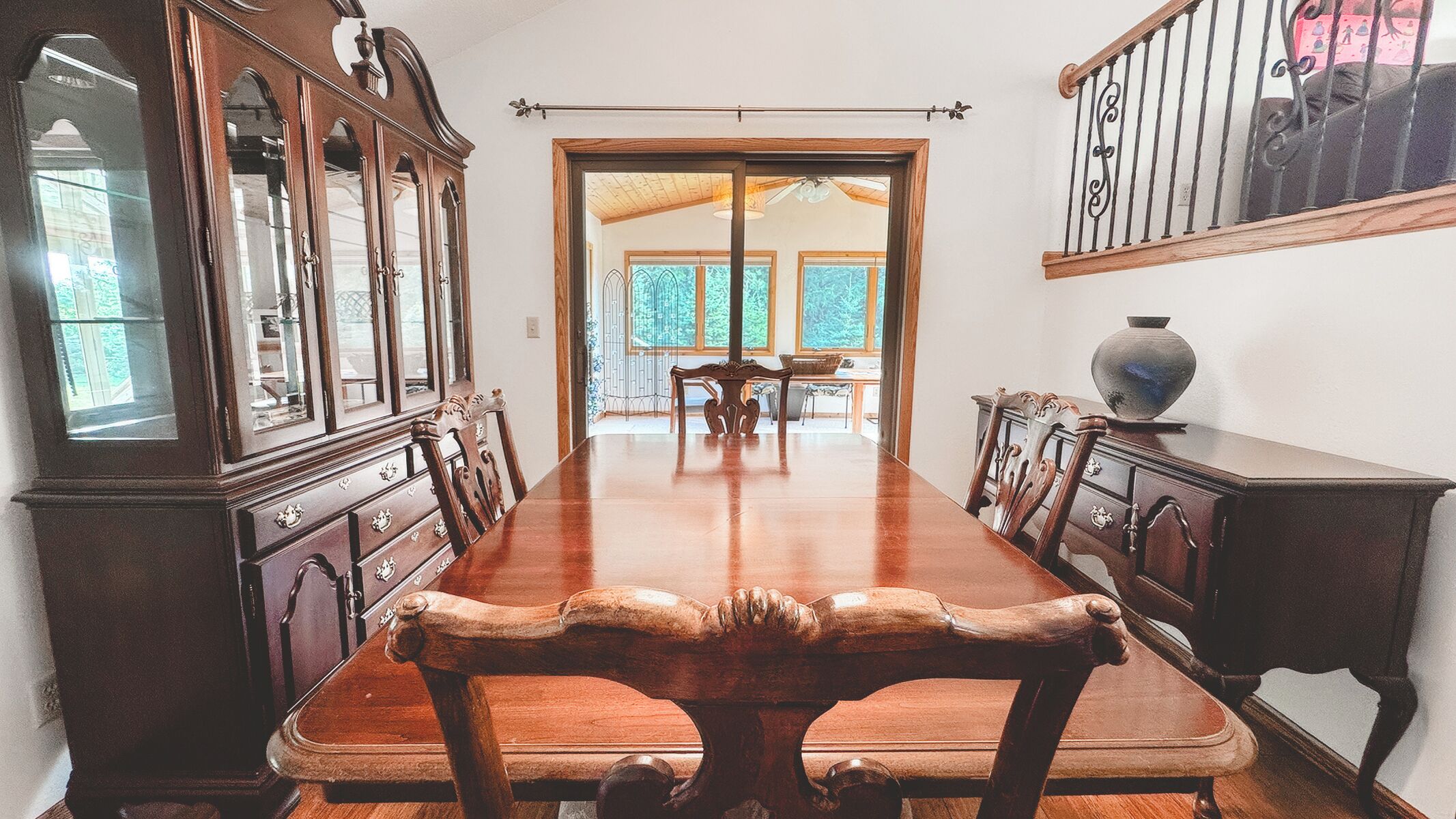 Una larga mesa de comedor de madera con sillas ornamentadas, flanqueada por armarios de madera con puertas de vidrio, en una habitación con vistas al exterior.