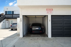 Un garaje con un SUV negro estacionado en su interior, un edificio blanco con una puerta de garaje negra, y un letrero que indica 'Solo Estacionamiento para Inquilinos Asignados'.
