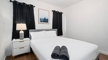A neatly made bed with white linens and two rolled towels, a white bedside table with a lamp, and black curtains on either side.