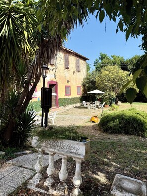 A courtyard with a stone bench, outdoor furniture, and a building with shutters. - Torvaianica, Zoomarine, Rome: Seaside Apartment with Shared Garden (Lazio)
