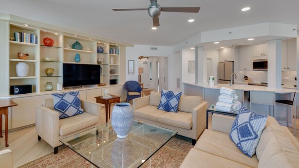 A modern living room with a flat-screen TV, two sofas with patterned cushions, a glass coffee table, and a ceiling fan. - 1617 Lands End Village | Best View on Captiva! (Captiva)