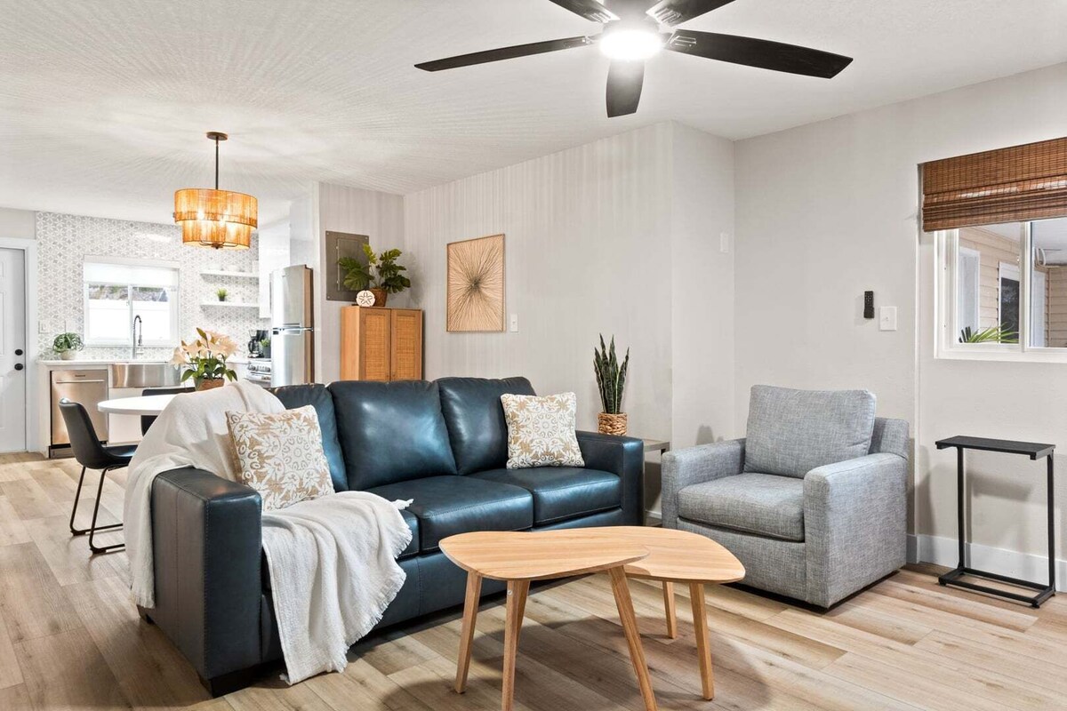 A modern living room with a black leather sofa, a grey armchair, and a wooden coffee table. There is a ceiling fan, a kitchen in the background, and a window with blinds.