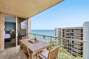 Ein Balkon mit einem Holztisch und Stühlen, mit Blick auf das klare, blaue Meer und ein mehrstöckiges Gebäude.