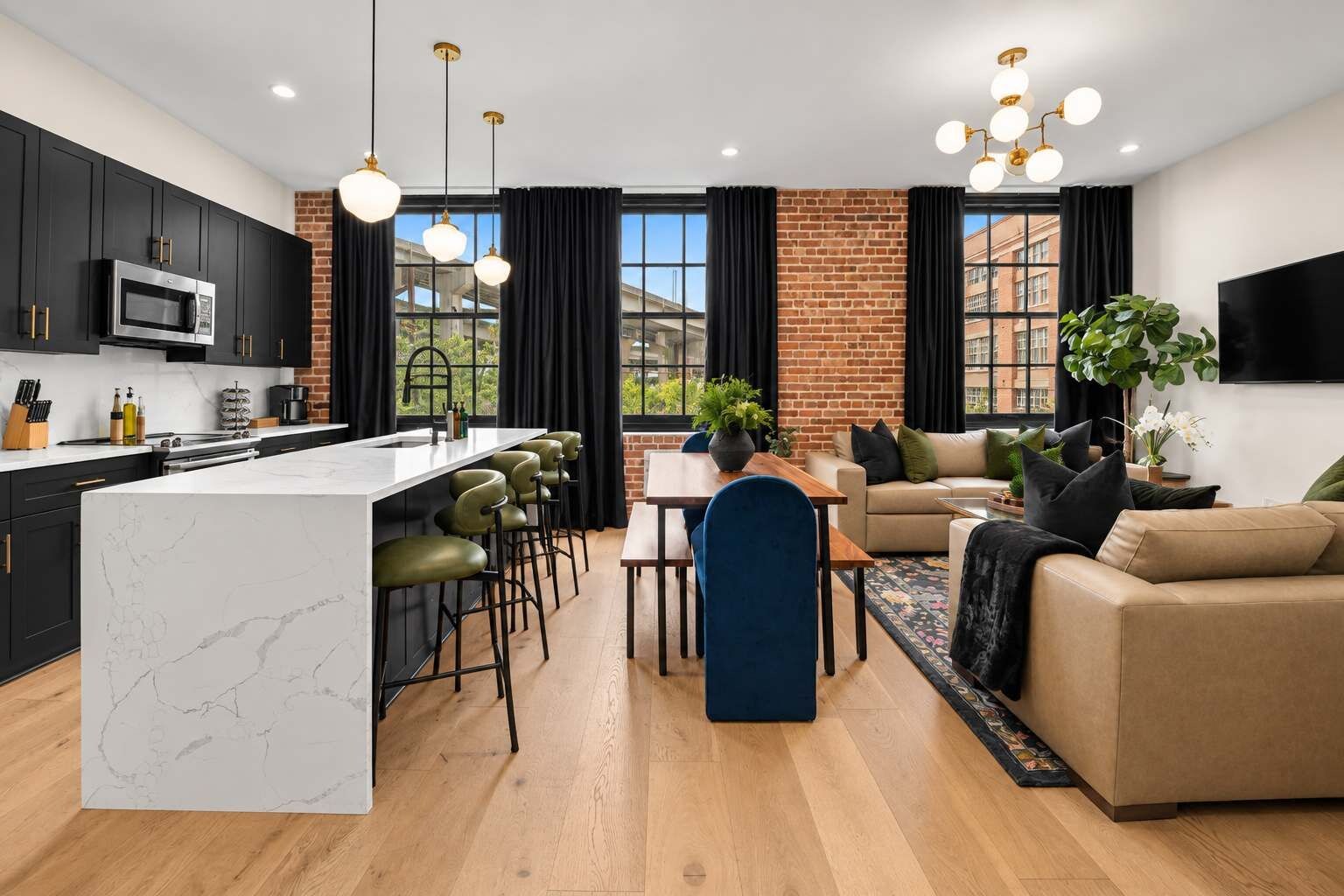 A modern kitchen with a central island, bar stools, a dining area with a table and chairs, a sofa, and a TV mounted on the wall.