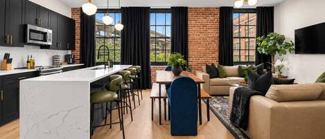 Een moderne keuken met een centraal eiland, barstoelen, een eethoek met tafel en stoelen, een bank en een aan de muur bevestigde televisie.