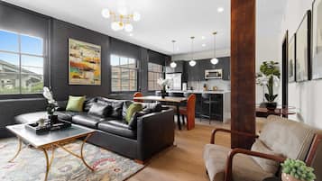 A modern living room with a black leather sofa, a coffee table, and a rug. There is a kitchen area with a dining table and chairs.