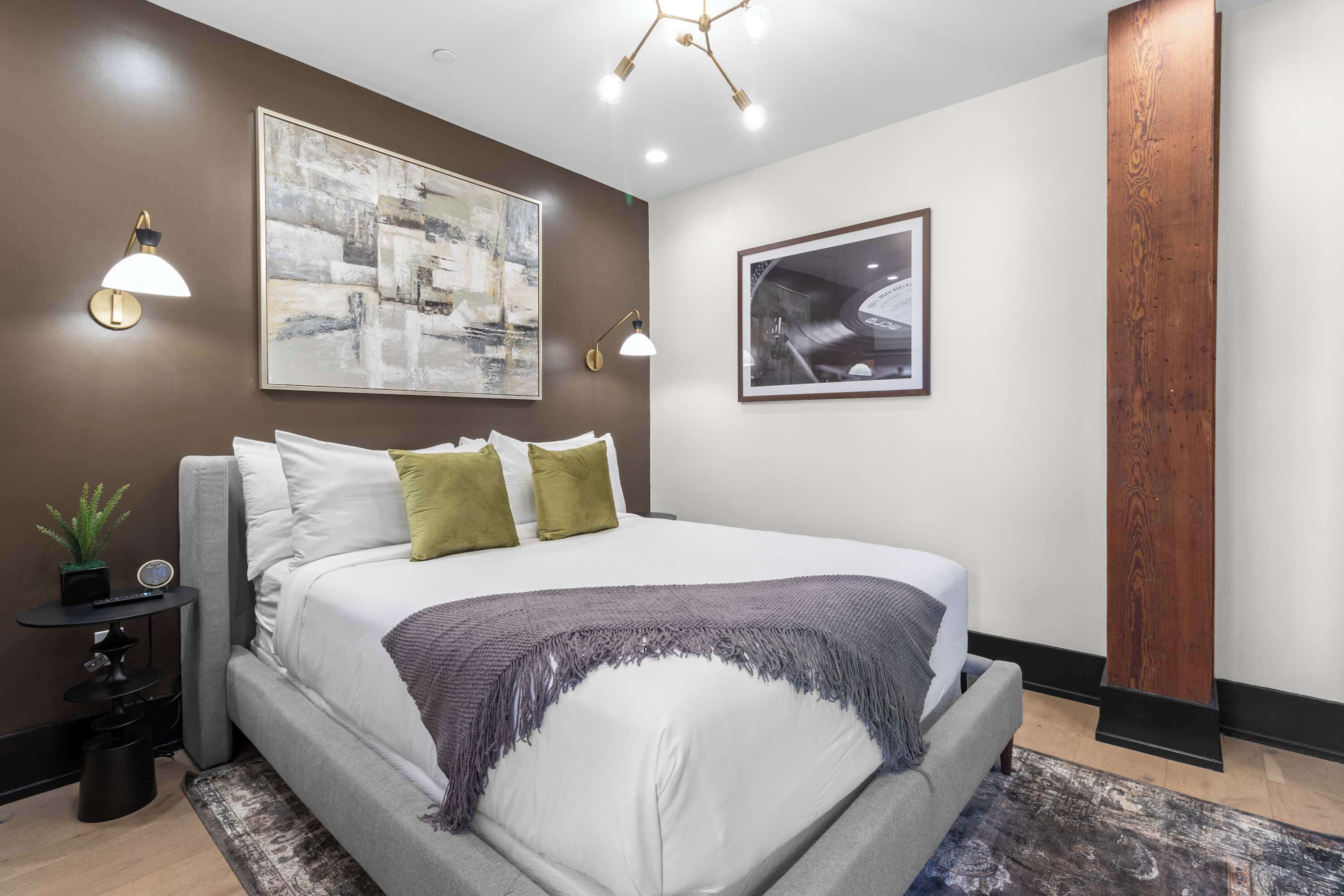 Ein modernes Schlafzimmer mit einem großen Bett, zwei Wandleuchten, einem gerahmten abstrakten Gemälde und einer hohen Holzsäule.