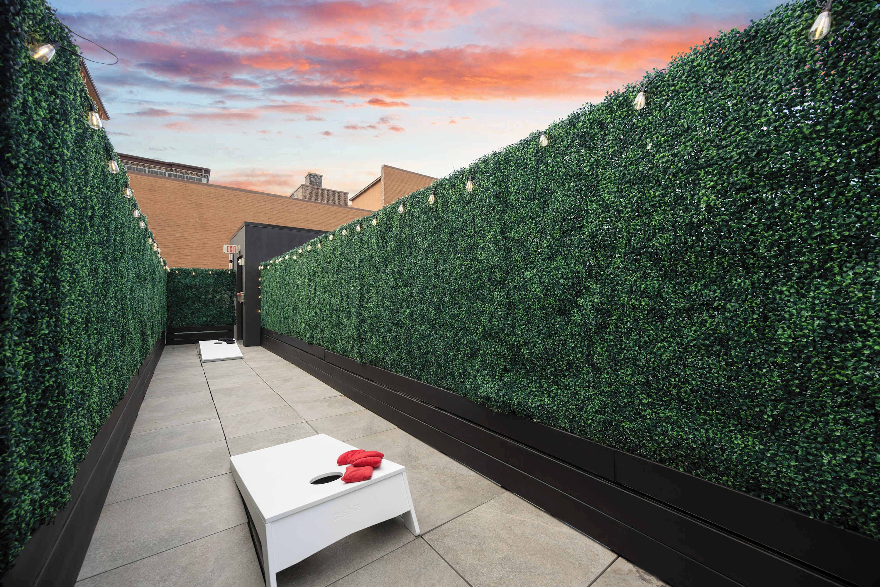 A narrow outdoor corridor with a white table and red cushions, flanked by tall green hedges, leading to an exit sign.