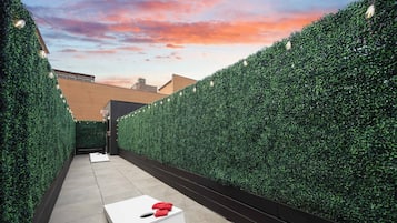 A narrow outdoor corridor with a white table and red cushions, flanked by tall green hedges, leading to an exit sign.