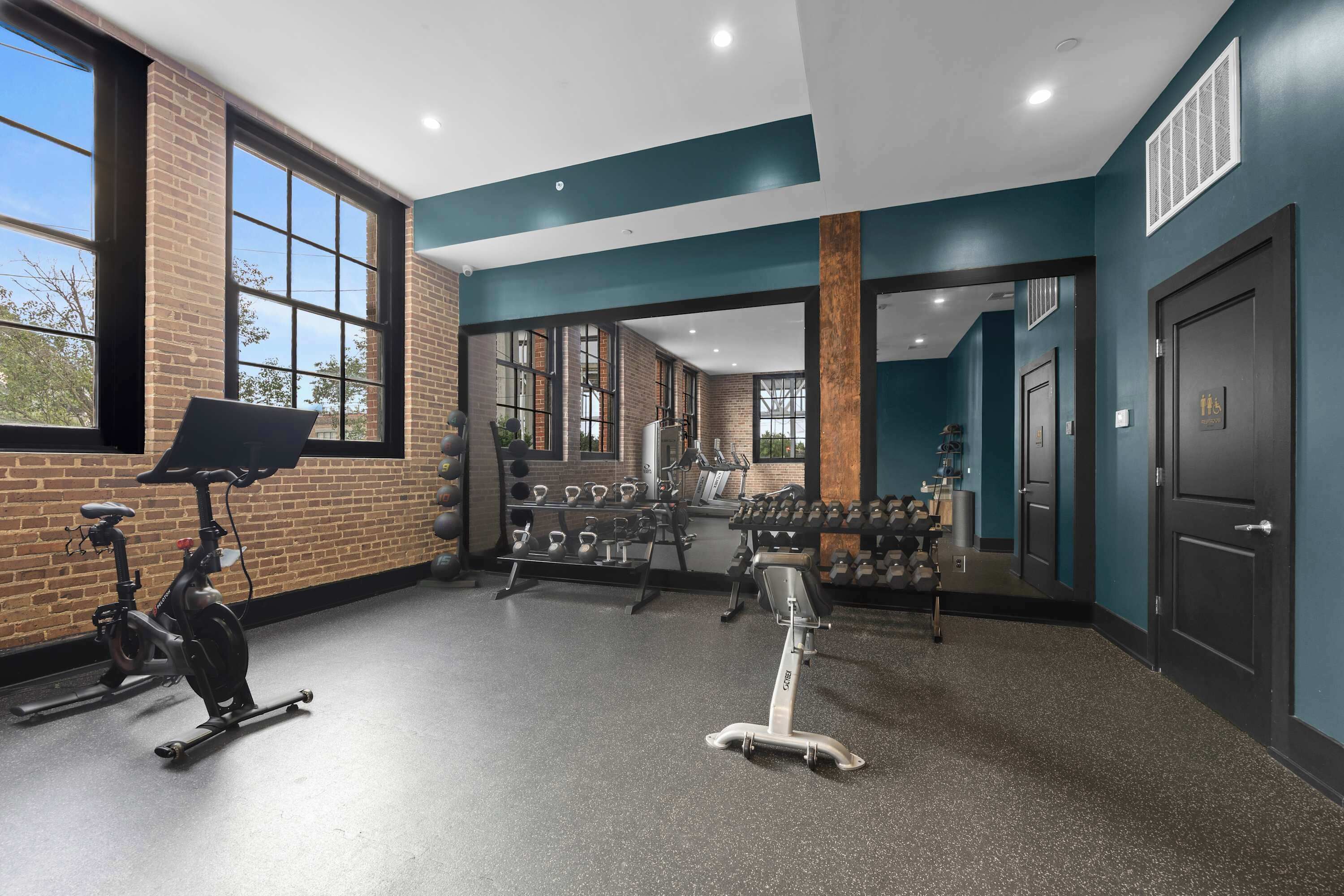 Ein Fitnessstudio mit Trainingsgeräten, großen Fenstern und einer Ziegelwand.