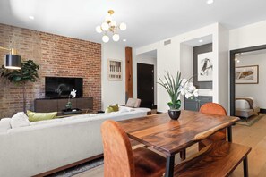 A modern living room with a brick wall, a flat-screen TV, a wooden dining table, and a sofa. - Connecting 6 Bedroom W/ 2 Living Rooms Sleeps 16 (New Orleans)