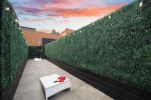 A narrow outdoor corridor with a white table and red cushions, flanked by tall green hedges, leading to an exit sign. - Downtown Hotel 2 King Bed Suite | Gym | Rooftop (New Orleans)