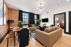 A modern hotel room with a brick accent wall, a dining area with a wooden table and chairs, a sofa, a flat-screen TV, and a large window with curtains. - Downtown Hotel 2 King Bed Suite | Gym | Rooftop (New Orleans)