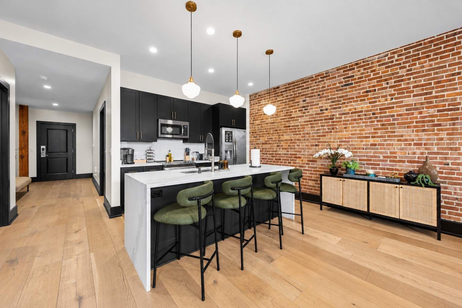 Une cuisine moderne avec un mur en briques, des meubles noirs, un îlot central blanc avec des tabourets de bar et des luminaires suspendus.
