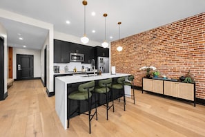Une cuisine moderne avec un mur en briques, des meubles noirs, un îlot central blanc avec des tabourets de bar et des luminaires suspendus.