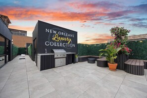 Eine Außenpatio-Terrasse mit Grillstation und Sitzgelegenheiten, einem Schild mit der Aufschrift 'NEW ORLEANS Luxury Collection' und einem Sonnenuntergang als Hintergrund.