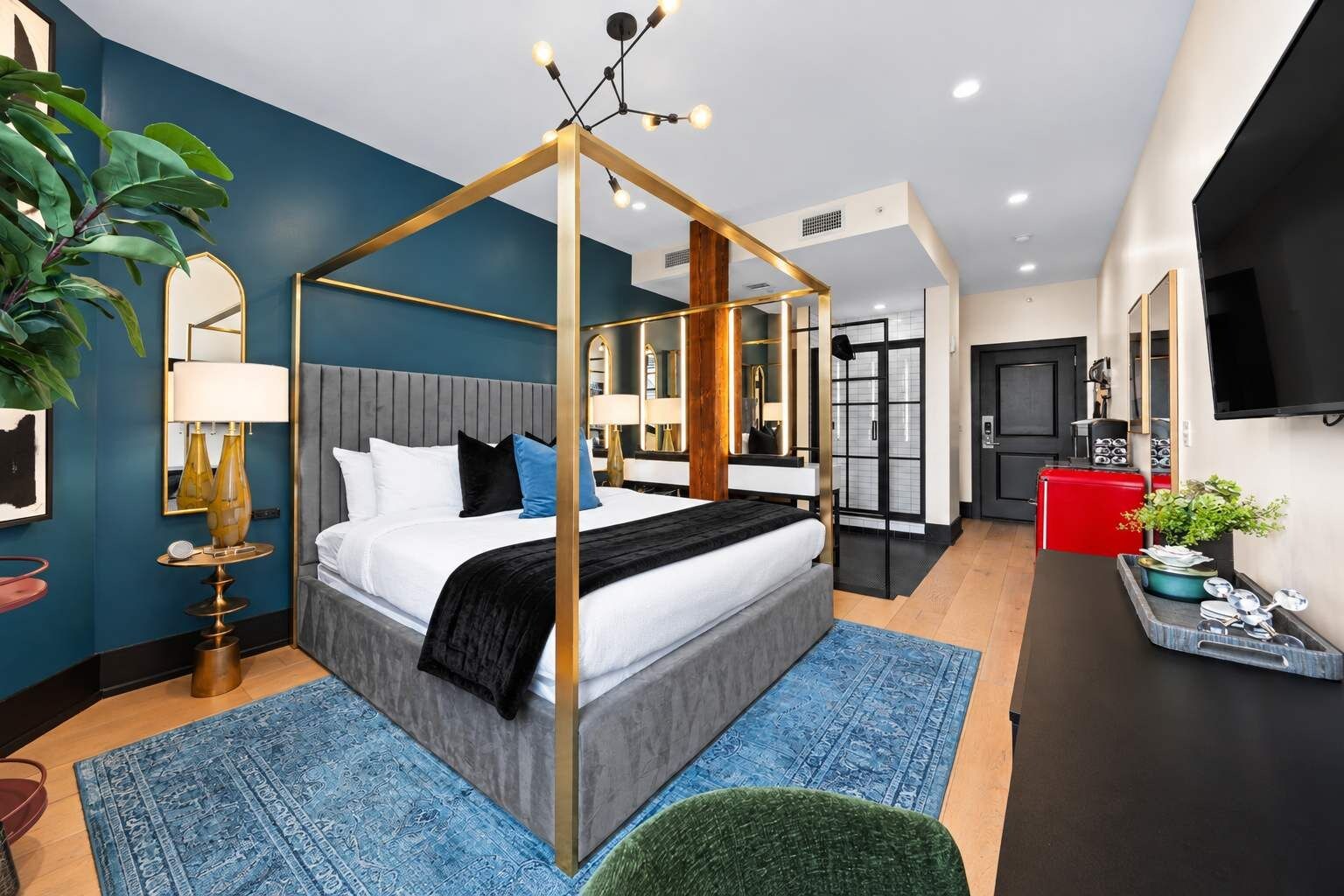 Ein modernes Hotelzimmer mit Himmelbett, Flachbildfernseher, rotem Koffer und einer großen Topfpflanze.