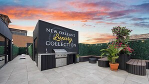 An outdoor patio area with a grill station and seating, a sign reading 'NEW ORLEANS Luxury Collection,' and a backdrop of a sunset sky. - All New | King bed | Gym | Rooftop (New Orleans)