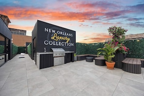 Eine Außenpatio-Terrasse mit Grillstation und Sitzgelegenheiten, einem Schild mit der Aufschrift 'NEW ORLEANS Luxury Collection' und einem Sonnenuntergang als Hintergrund.