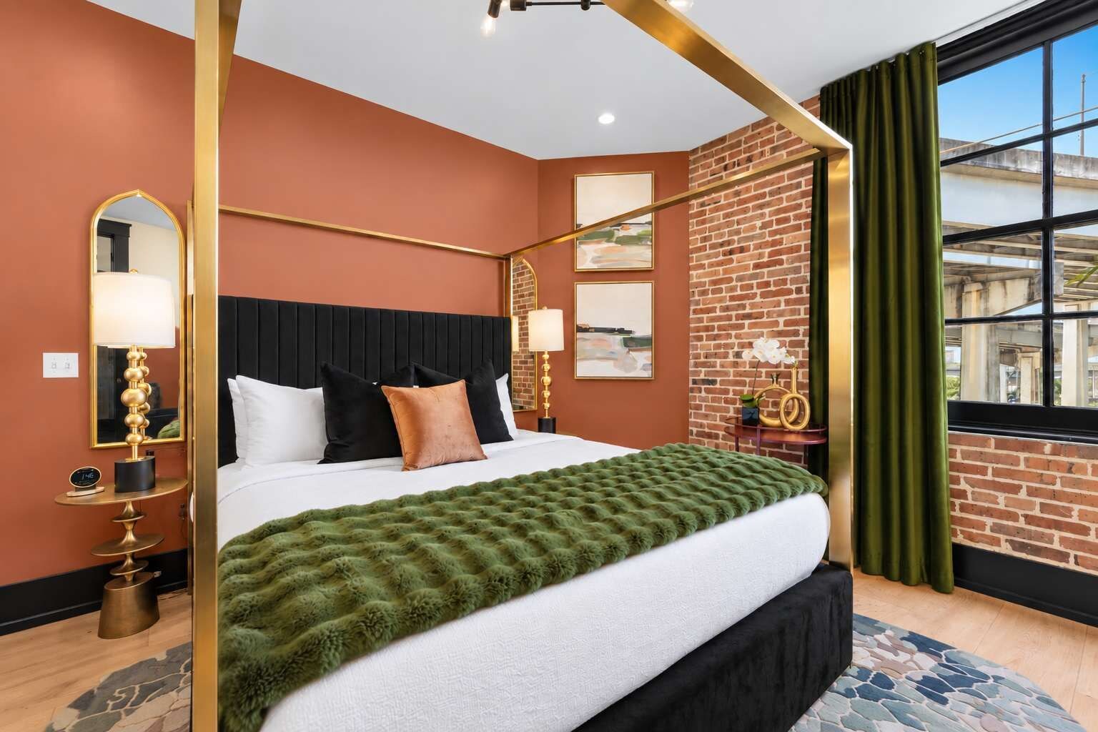 A hotel room with a four-poster bed, a brick accent wall, a large window with curtains, and a green throw blanket.