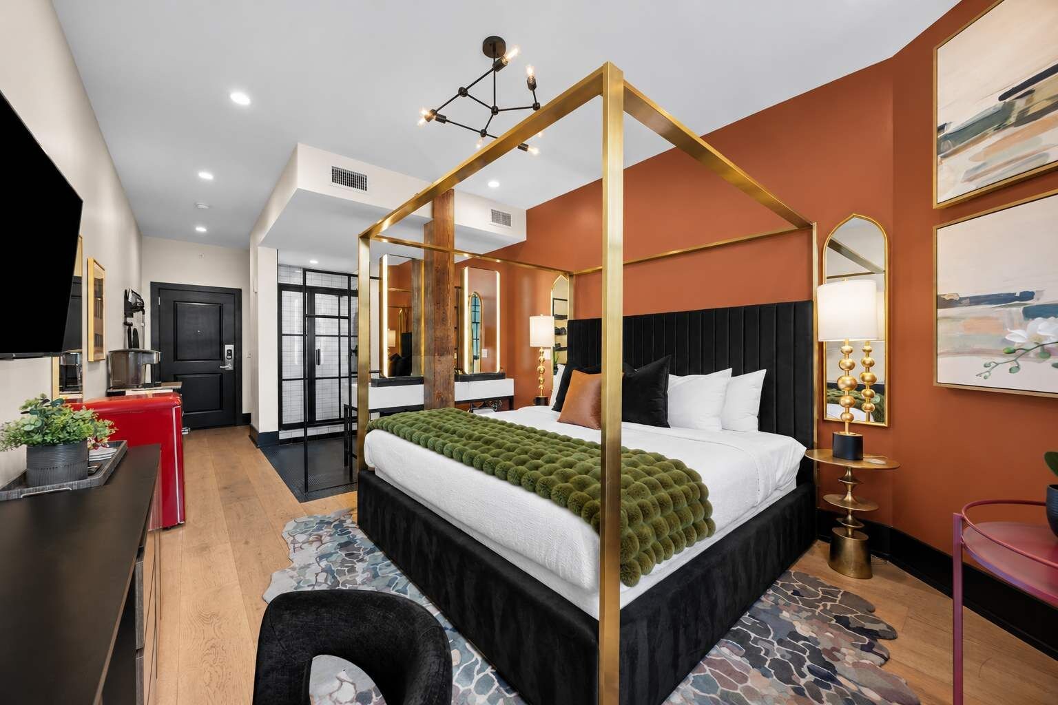 A modern hotel room with a four-poster bed, a desk with a chair, a small table, and a red cabinet.