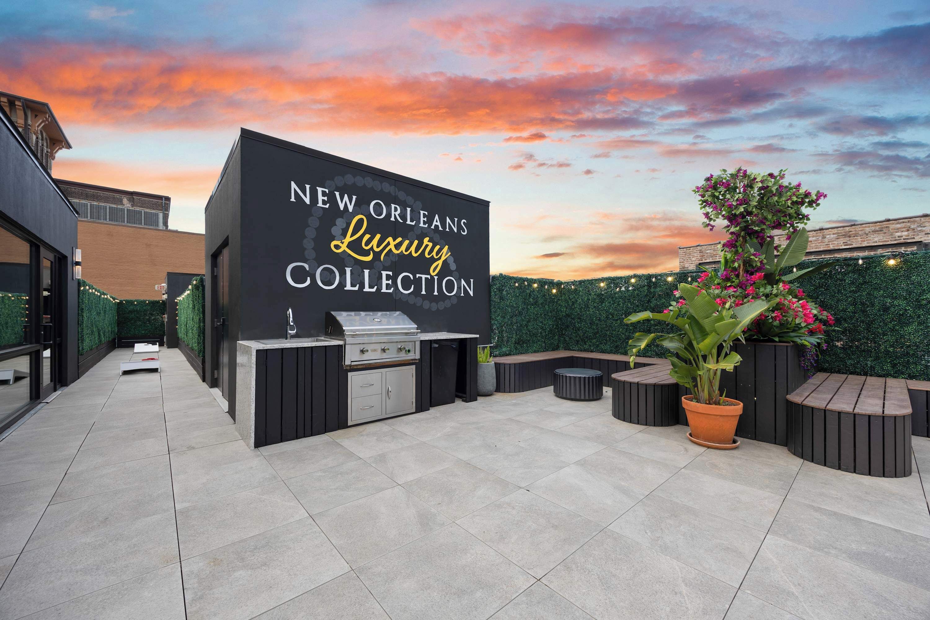 Un espace extérieur aménagé en terrasse, avec un coin barbecue et des sièges, une enseigne indiquant « NEW ORLEANS, Collection de luxe », et un ciel au coucher du soleil en arrière-plan.