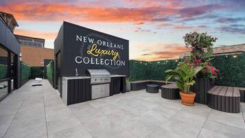 Un espace extérieur aménagé en terrasse, avec un coin barbecue et des sièges, une enseigne indiquant « NEW ORLEANS, Collection de luxe », et un ciel au coucher du soleil en arrière-plan.