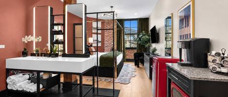 A modern hotel room with a large bed, a separate sitting area, a kitchenette, and a bathroom with a large mirror and sink.