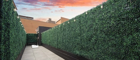 A narrow outdoor corridor with a white table and red cushions, flanked by tall green hedges, leading to an exit sign.