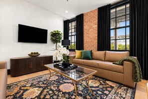 A modern living room with a leather sofa, a glass coffee table, a TV mounted on the wall, a potted plant, and a large window with curtains. - Sleeps 10 | Luxury 4BR Downtown Hotel (New Orleans)