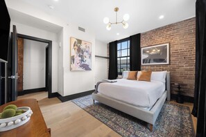 A modern bedroom with a large bed, a wooden side table, a painting, and a brick wall. - Sleeps 10 | Luxury 4BR Downtown (New Orleans)