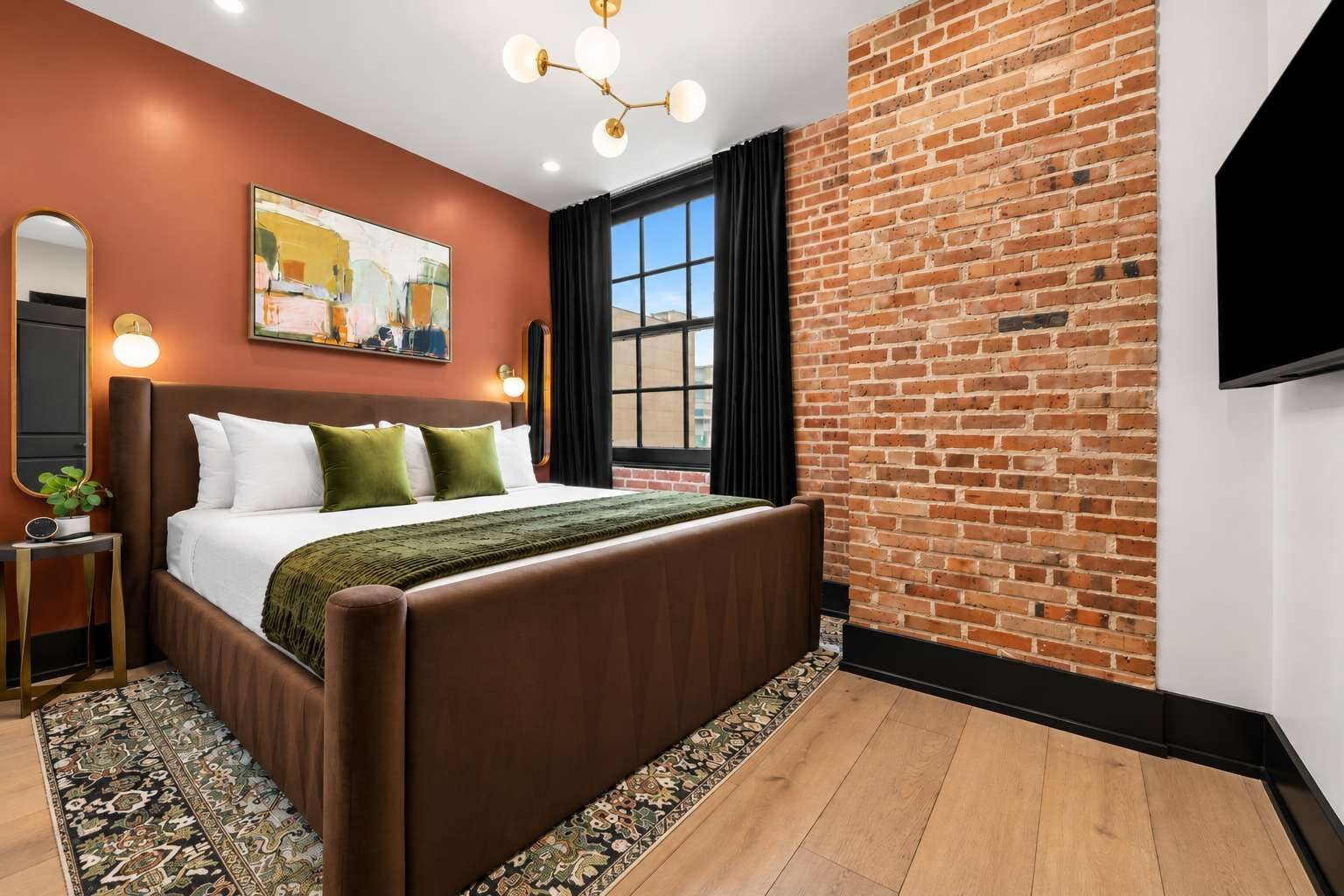 Un dormitorio moderno con pared de ladrillo, una cama grande, un televisor y una ventana con cortinas.