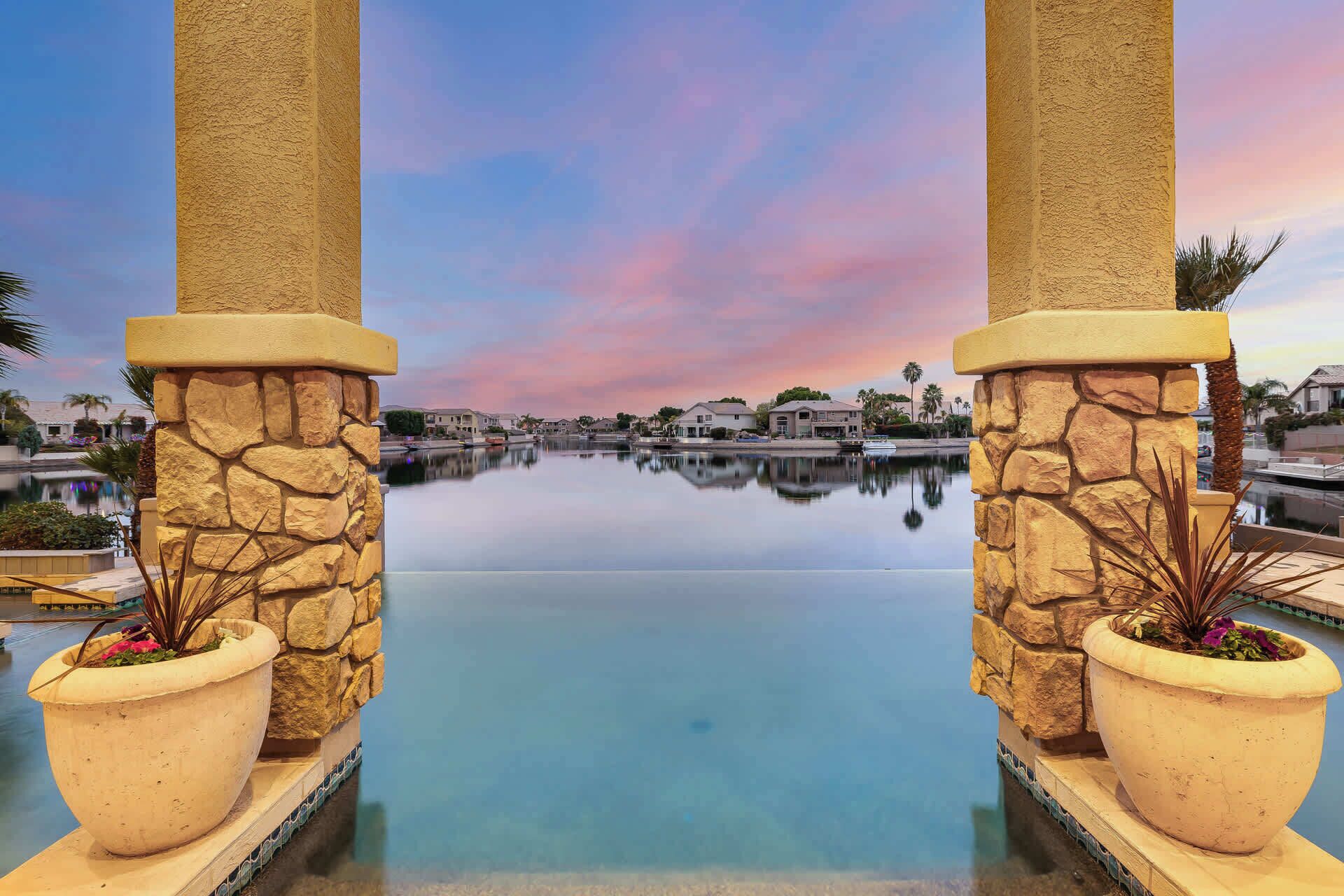 A pool with stone pillars and potted plants, surrounded by a calm water body and a sunset sky.