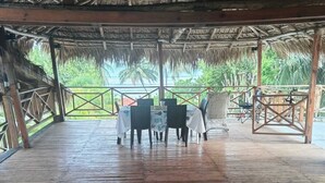 Una terraza de madera al aire libre con techo de paja, rodeada por una barandilla metálica, con vistas a un paisaje tropical.