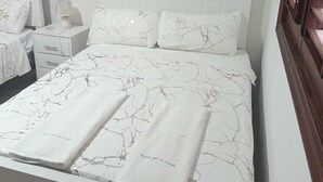 Una cama bien hecha con cabecera blanca y dos almohadas, una mesita de noche con una lámpara y un persiano de madera.