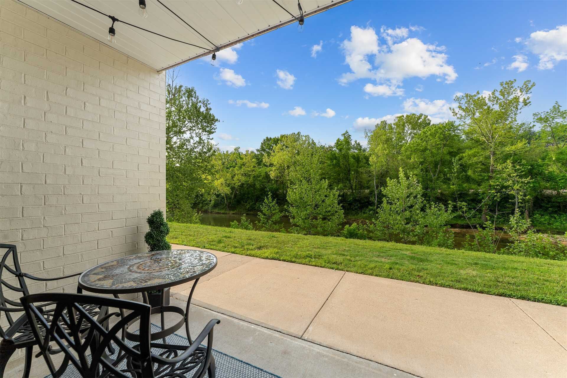 A patio with a table and chairs, a brick wall, and a view of a grassy area with trees and a body of water.