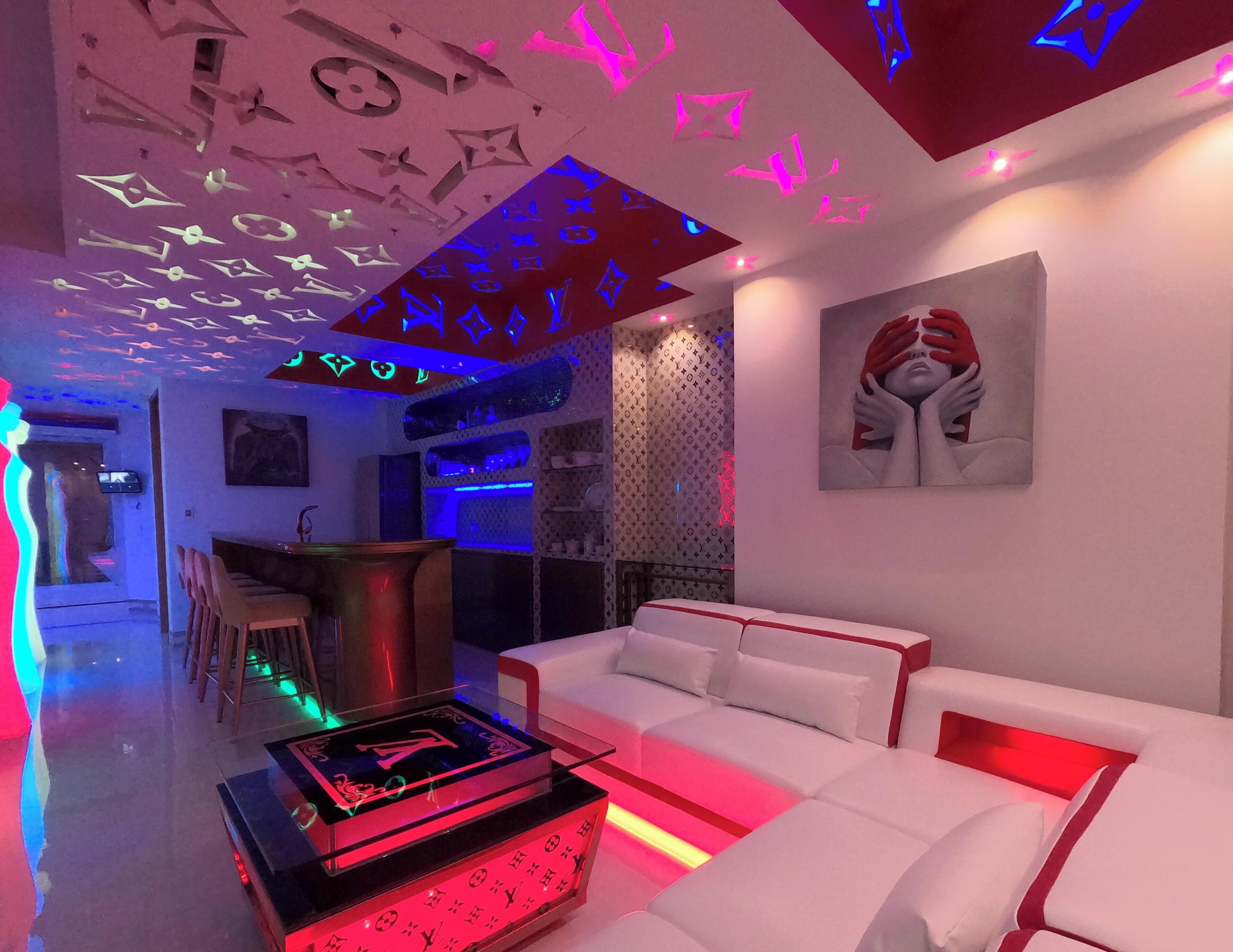 Una sala de estar moderna con un sofá blanco, una mesa de centro de vidrio con diseño Louis Vuitton, y una zona de bar con iluminación azul.
