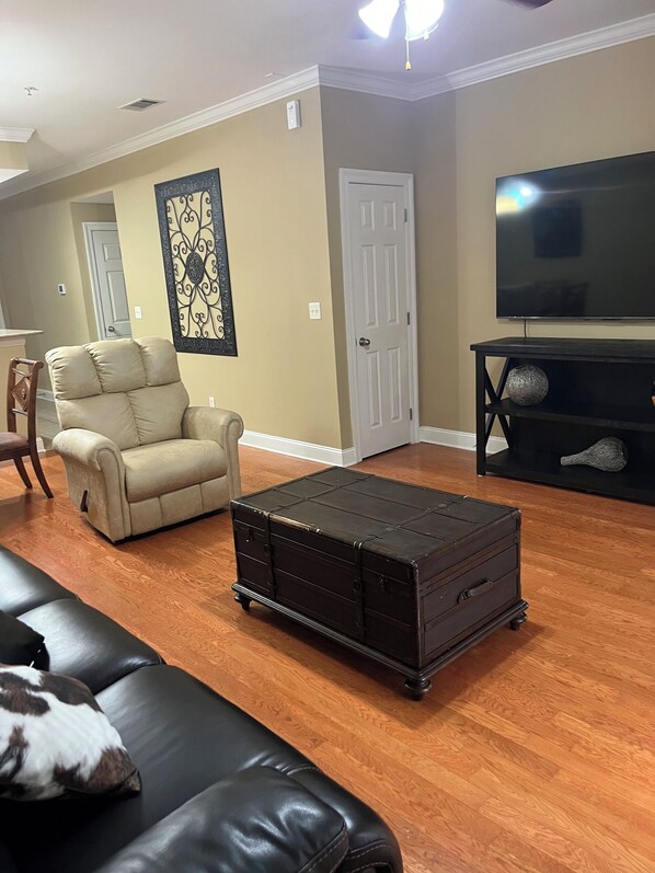 A living room with a black leather sofa, a beige armchair, a wooden coffee table, a flat-screen TV on a black stand, and a decorative wall piece.